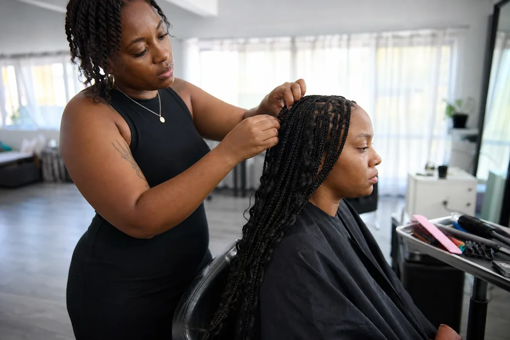 A stylist finishing a long protective style with hair extensions on a seated client in a small studio